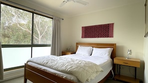 A neatly made bed with white linens, a wooden headboard, and a bedside table with a lamp. A window with curtains allows natural light into the room. - A charming family-friendly retreat nestled in the tranquil heart of Venus Bay (Venus Bay)