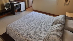 Een slaapkamer met een bed, een bureau, een stoel, een televisie, een klok en een raam met een plant.