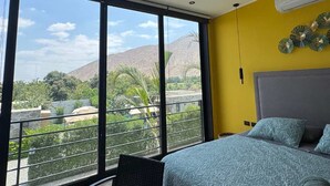 Een slaapkamer met een groot raam, een bed, een stoel en uitzicht op een berg en groen.