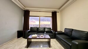 A modern living room with a leather sofa, a glass coffee table, and a view of a cityscape through large windows. - Margarita Hotel Amman (Amman)