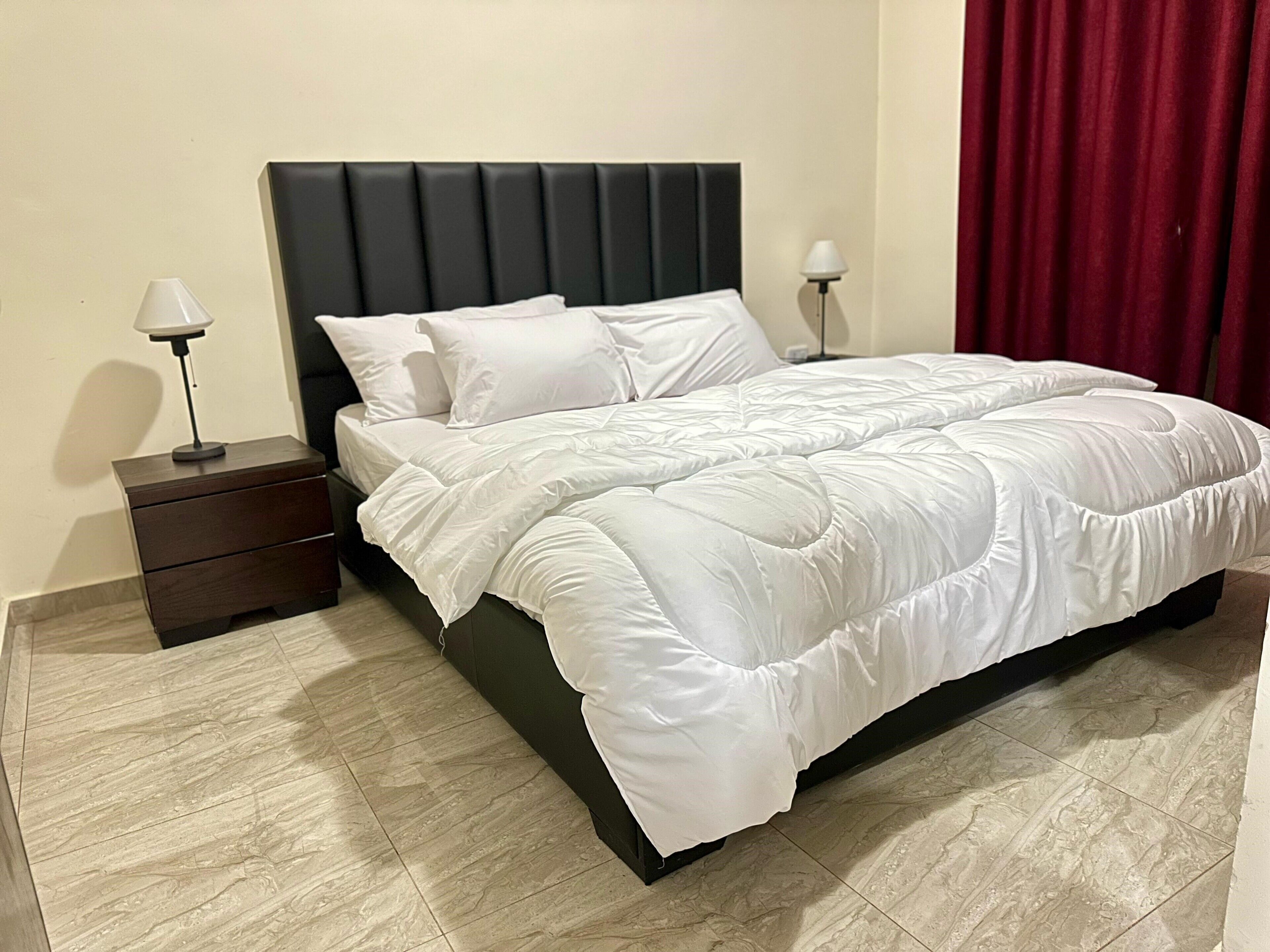 A neatly made bed with white bedding, a dark headboard, and a bedside table with a lamp.