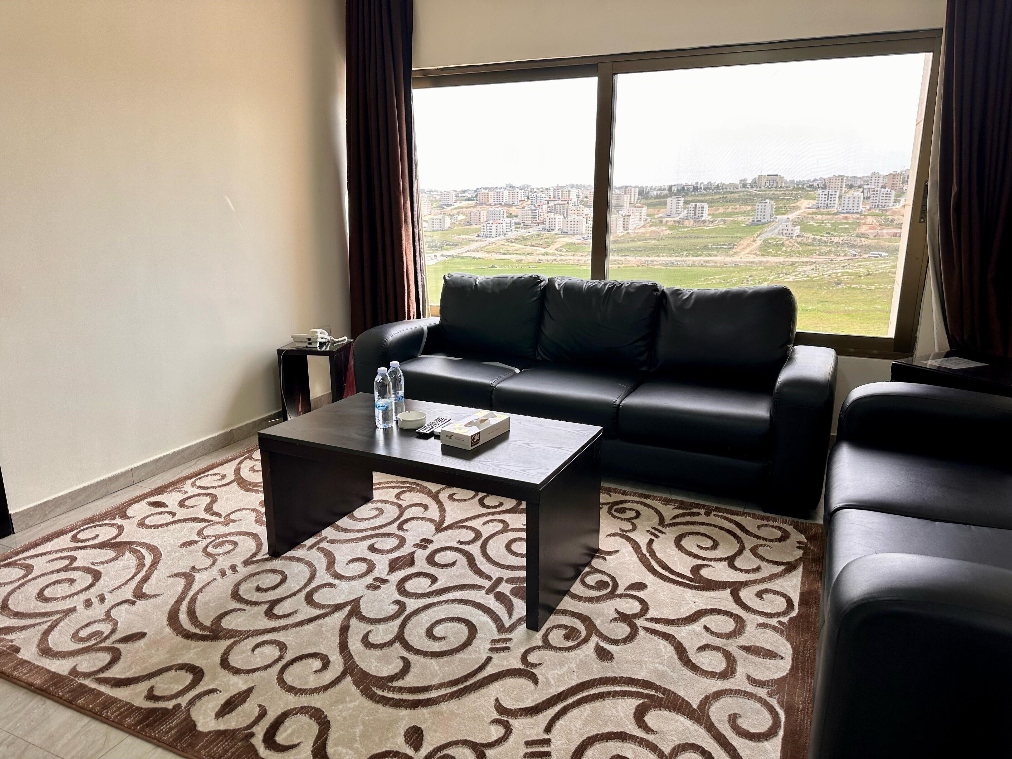 A living room with a black leather sofa, a coffee table, and a view of a cityscape through large windows.