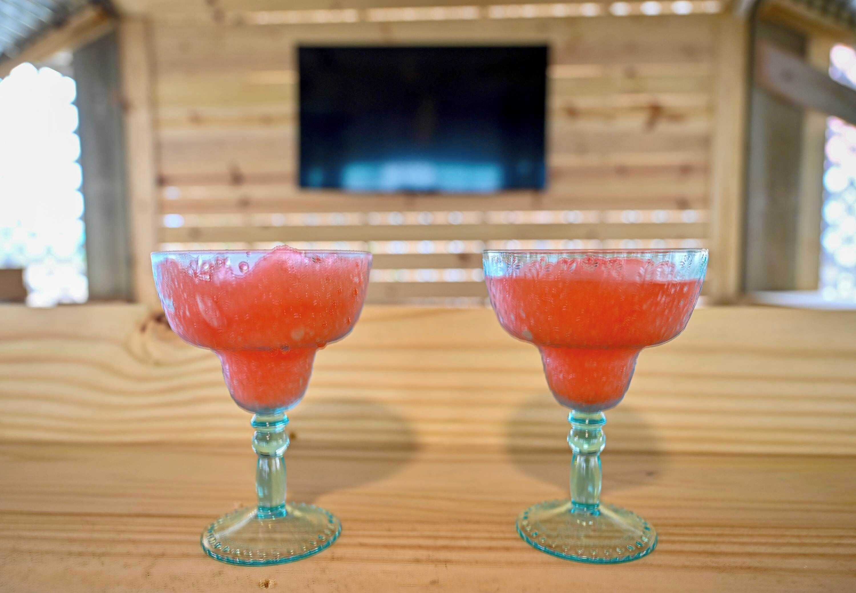 Dos cócteles en vasos de tallo sobre una mesa de madera, con una pared de madera y un televisor de pantalla plana al fondo.