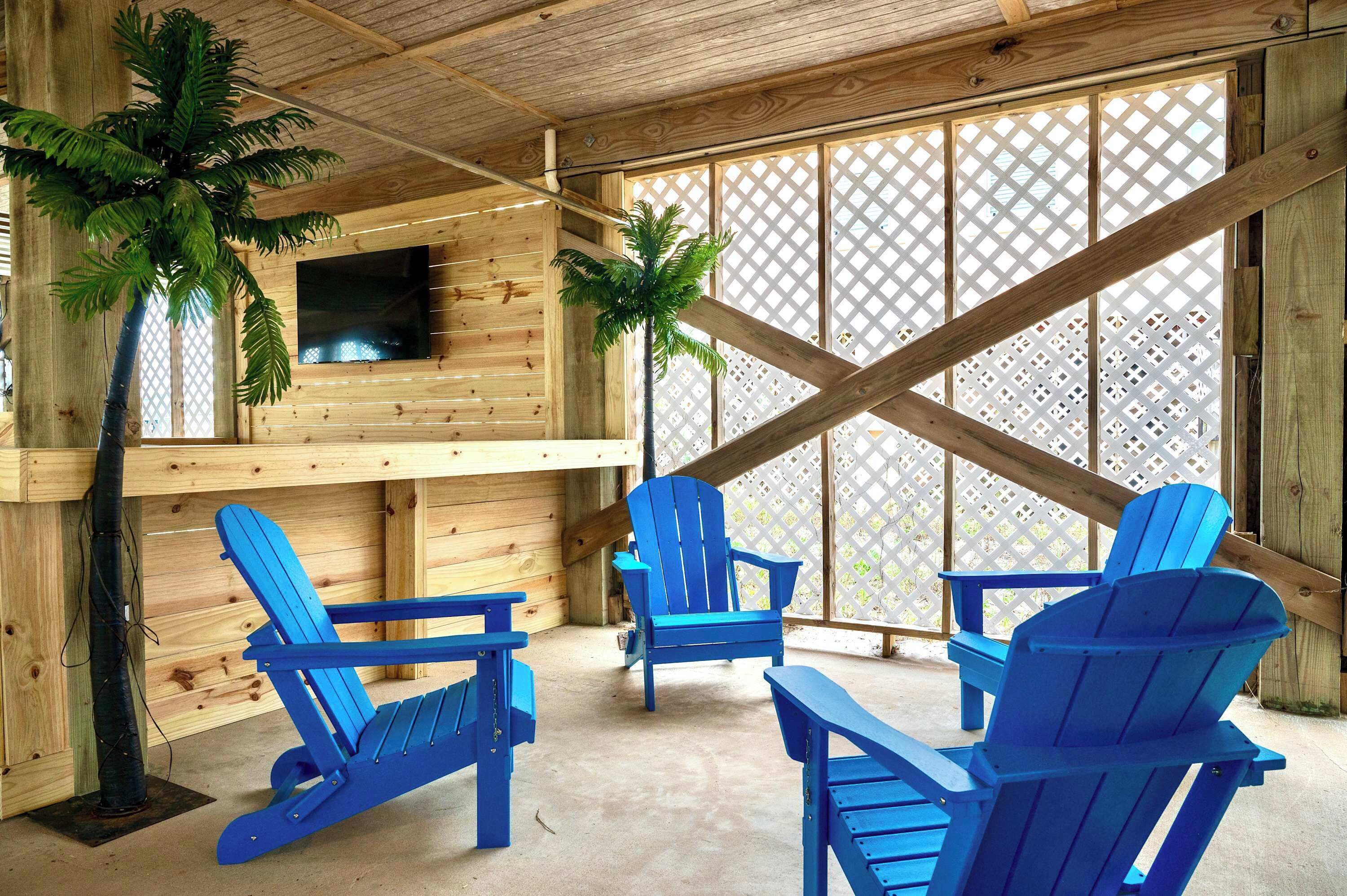 A wooden interior with blue Adirondack chairs, a TV mounted on the wall, and two potted palm trees.