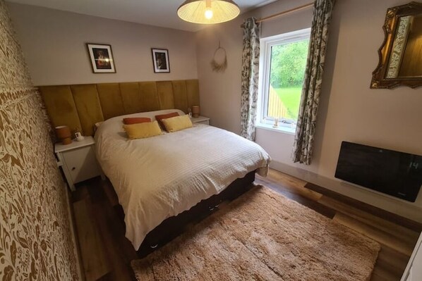 Un dormitorio con cama, mesitas de noche, televisor y ventana con cortinas.