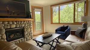 Un salon avec une cheminée en pierre, un fauteuil bleu, un canapé beige et une grande fenêtre donnant sur la forêt.