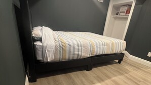 Ett rum med en säng placerad mot en mörk vägg, ett ljust trägolv och ett vitt tak med infälld belysning.