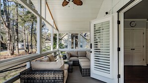 Une véranda couverte avec des sièges d’extérieur, une terrasse en bois et une vue sur les arbres et les maisons environnantes.
