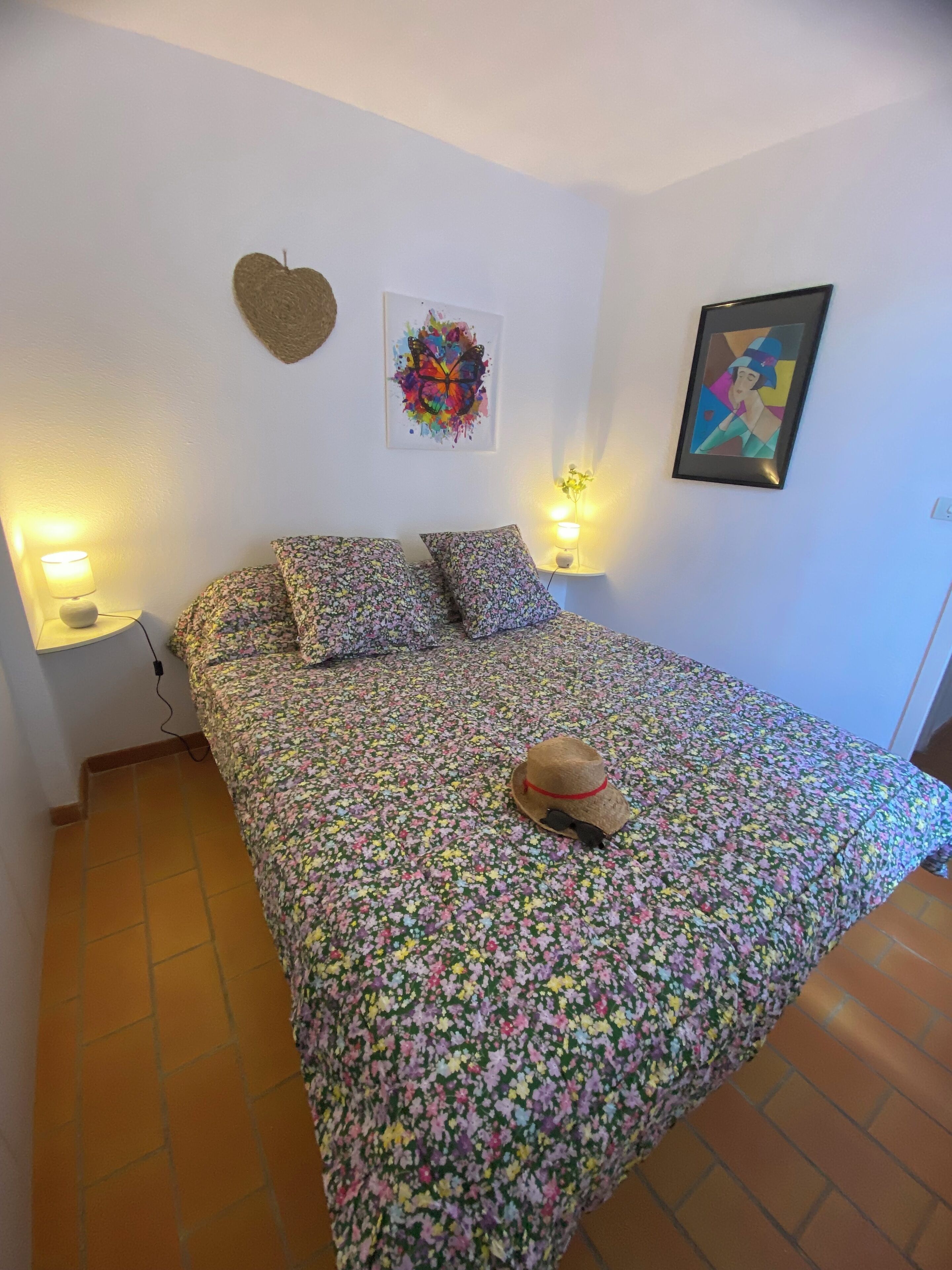 Ein Schlafzimmer mit geblumter Bettdecke, zwei Kissen, einem Hut, einer herzförmigen Dekoration und zwei Wandlampen.