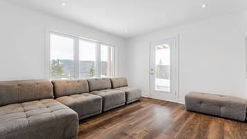 A modern living room with a sectional sofa, a small ottoman, and large windows offering a view of the outdoors.