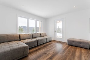 Ein modernes Wohnzimmer mit einem Ecksofa, einem kleinen Hocker und großen Fenstern mit Blick ins Freie.