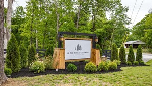 Une enseigne pour « In the Pines Cottages », avec un cadre en bois et un fond blanc, entourée d’un jardin aménagé avec différents arbustes et une clôture.