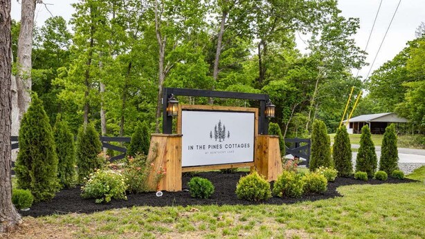 A sign for 'In the Pines Cottages' with a wooden frame and a white background, surrounded by a landscaped garden with various shrubs and a fence. - The Salt Shack ("In The Pines" at KY Lake) | 121 Pine Grove Lane, Dover, TN 37058 (Dover)