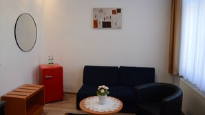 Ein modernes Wohnzimmer mit einem dunkelblauen Sofa, einem kleinen roten Kühlschrank, einer Holzbank, einem runden Couchtisch mit floraler Dekoration und einem an der Wand befestigten Spiegel.