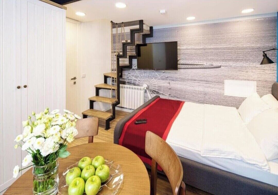 A modern hotel room with a bed, a TV mounted on the wall, a staircase, and a dining area with a table and chairs.