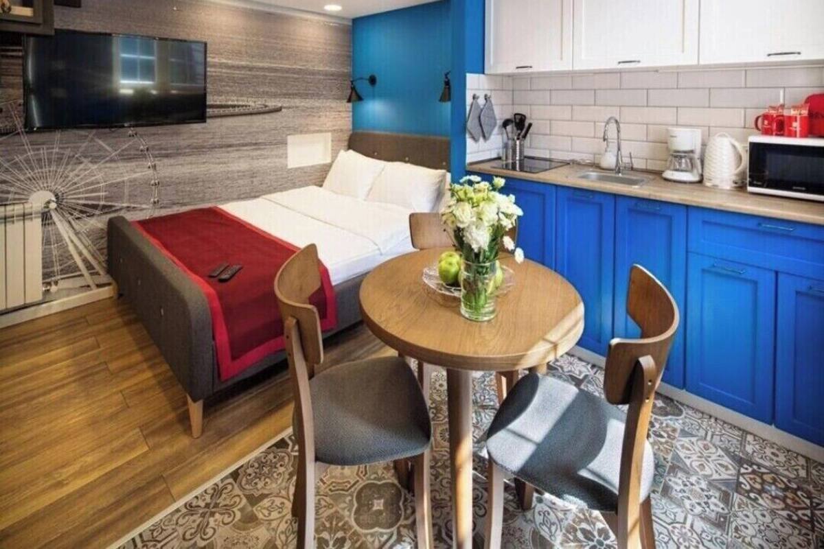 Une chambre d’hôtel compacte comprenant un lit, une petite table à manger avec des chaises, un téléviseur, un coin cuisine avec des placards bleus et un sol à motifs.