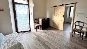 Una habitación con piso de madera, una cama con colcha estampada, una silla, un armario de madera y una puerta que da a otra habitación.