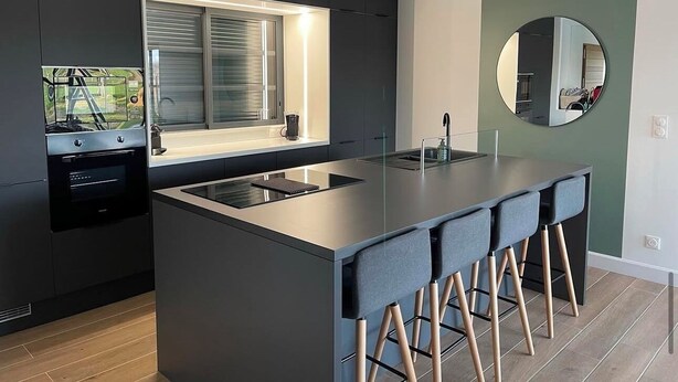 A modern kitchen with a central island, dark cabinetry, a window with blinds, and a round mirror on the wall. - Gîte Luna villa 6 persons with jacuzzi, children's playground and petanque area (Chaumont-sur-Loire)