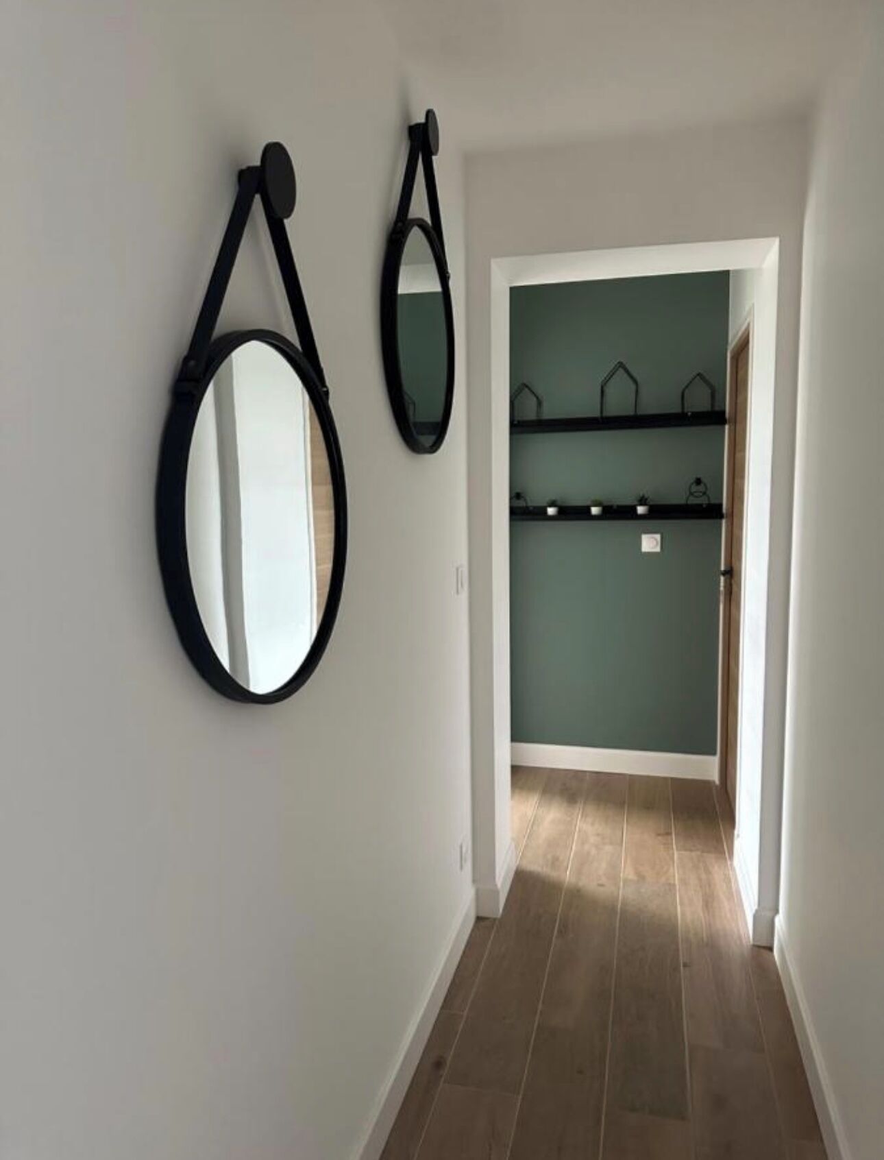 A hallway with a white wall, a black circular mirror, and a black rectangular mirror. There is a door leading to a room with a teal wall and shelves with decorative items.