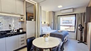 Ett kompakt hotellrum med ett litet pentry, en säng med utsikt, ett litet bord och en bordsfläkt.