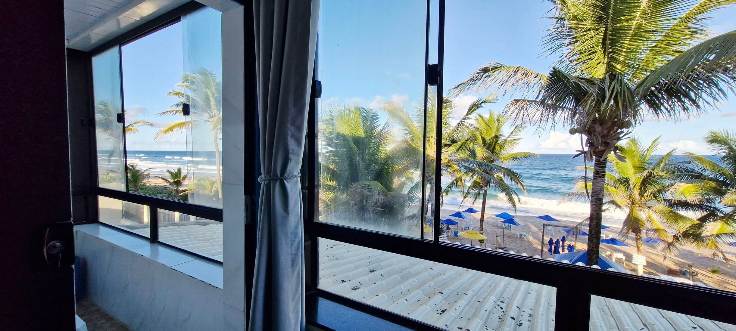 Vista desde una habitación con grandes ventanales que dan a una playa con palmeras y el océano.