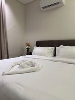 Una cama bien hecha con sábanas blancas y una toalla doblada, un cabecero oscuro, una mesita de noche con un florero y un aire acondicionado instalado en la pared.