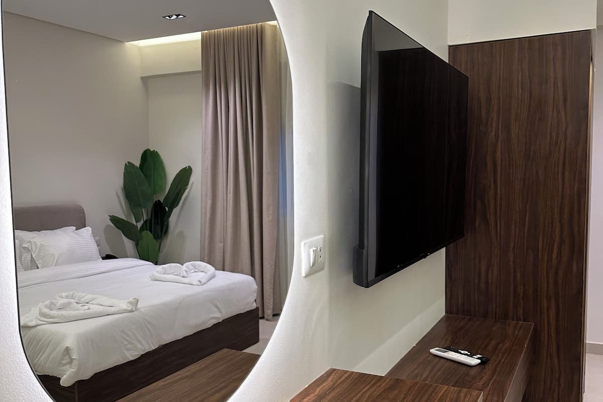 A modern hotel room with a large mirror, a flat-screen TV mounted on the wall, a wooden bench, and a bed with white linens.