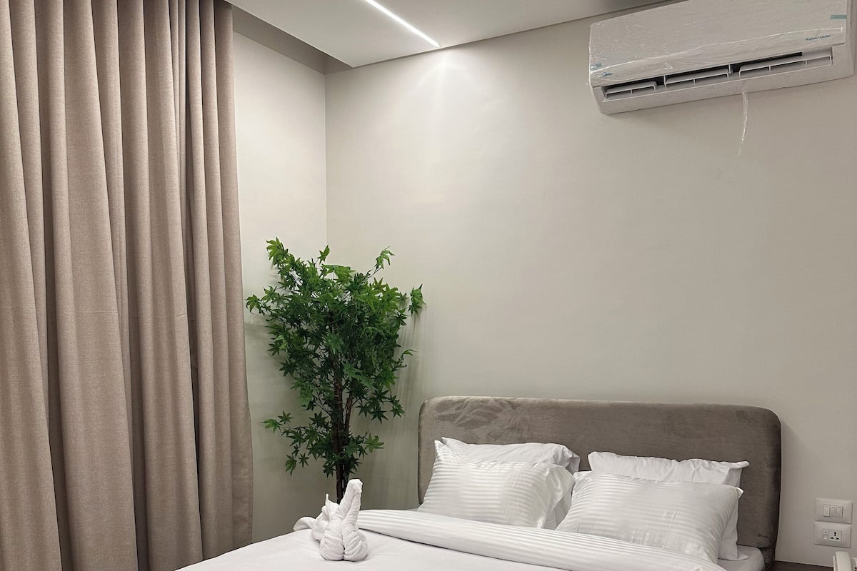 A neatly made bed with white linens, a dark wooden headboard, a potted plant, and an air conditioning unit on the wall.