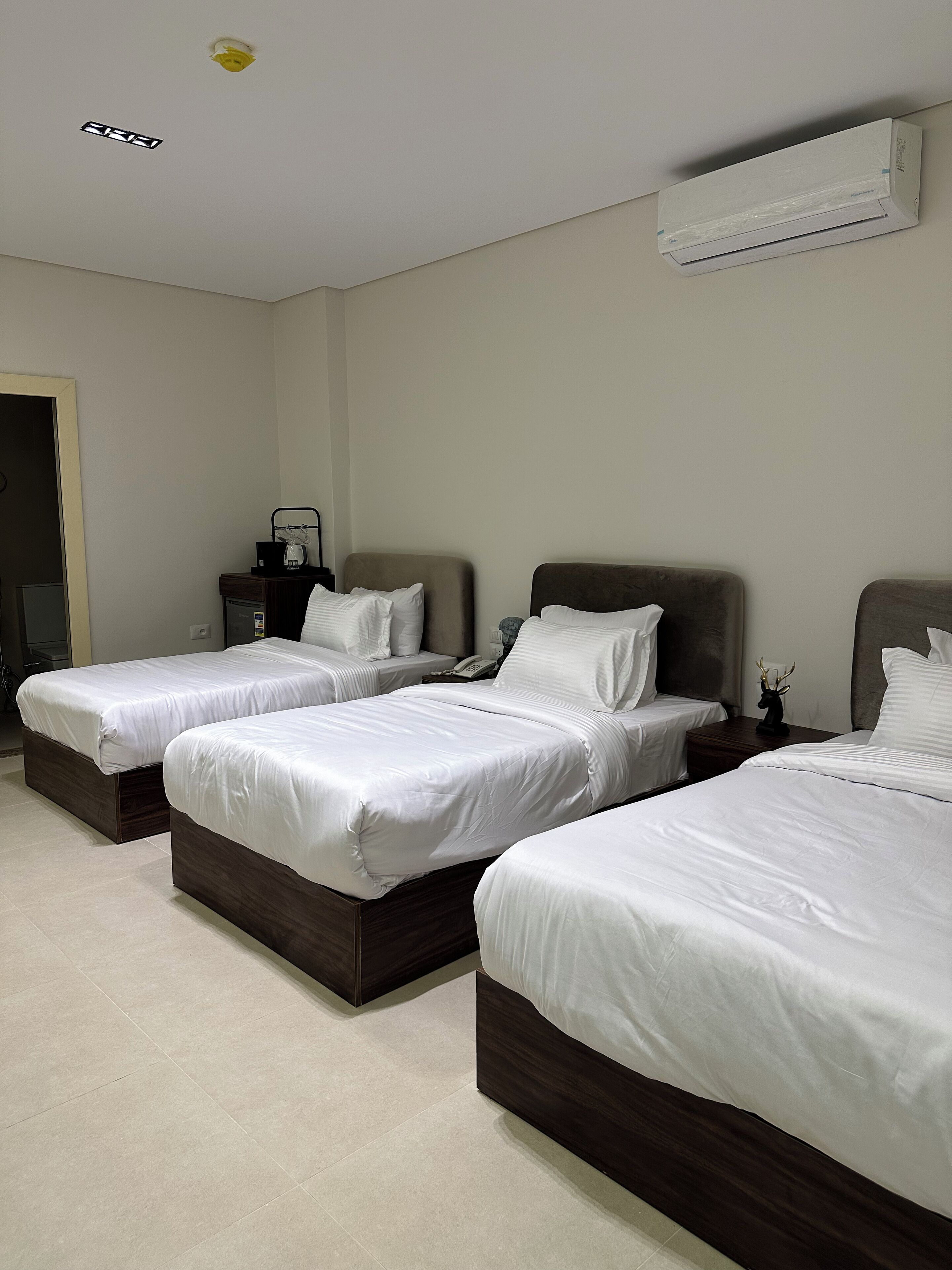 A hotel room with three single beds, a bedside table with a lamp, a phone, and a small decorative item.