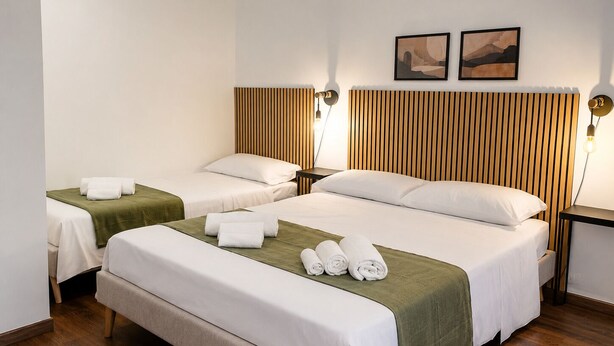 Habitación de hotel con tres camas, cabecera de madera, dos lámparas de pared y dos cuadros enmarcados.
