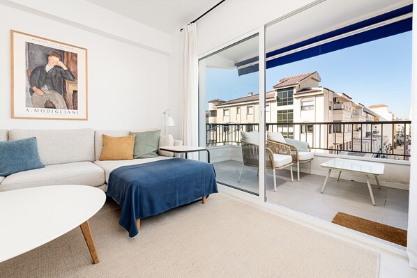 A modern bedroom with a sofa, a bed, a small table, and a balcony with a view of buildings.