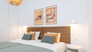 Ein modernes Schlafzimmer mit einem Holzkopfstück, einem Bett mit grün-beiger Bettwäsche, zwei gerahmten Kunstwerken an der Wand und einem kleinen weißen Nachttisch.