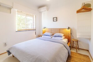 A bedroom with a bed, a bedside table, a hanging light, a window, and a wall-mounted air conditioner.