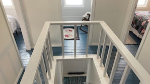 A staircase with white railings leading to a landing. A rocking chair is visible on the landing, and a bed with a floral bedspread is in the background.