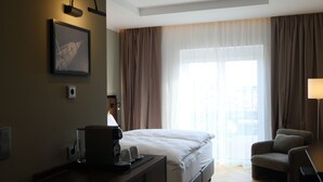 A hotel room with a bed, a desk with a coffee maker, a chair, and a large window with curtains. - UNICO Boutique Hotel (Hochheim am Main)