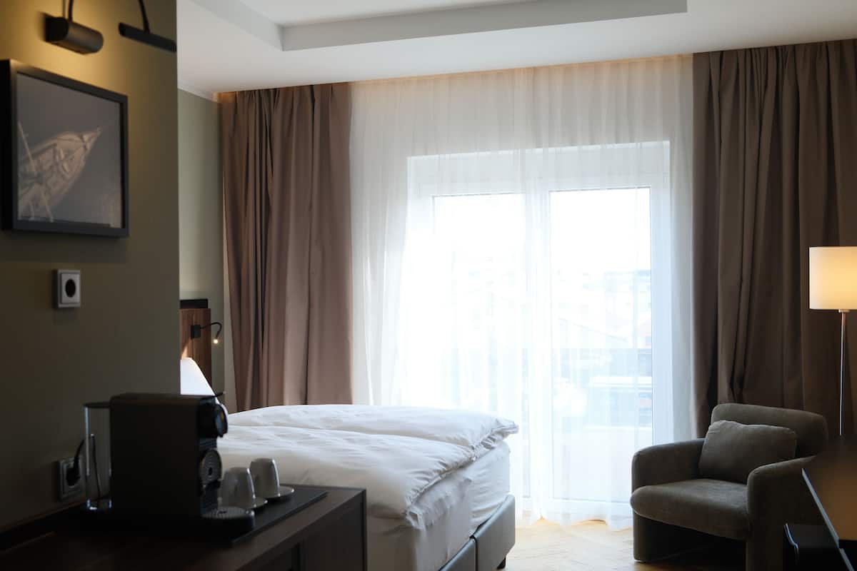 Hotellihuone, jossa on sänky, työpöytä, kahvinkeitin, tuoli ja suuri ikkuna, jossa on verhot.