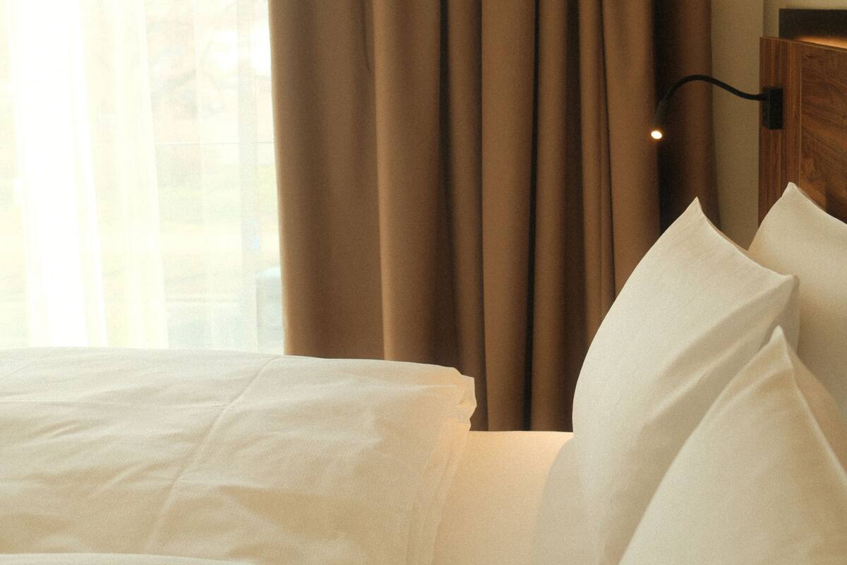Hotellihuone, jossa on sänky, verhot ja yöpöytä, jossa on lamppu.