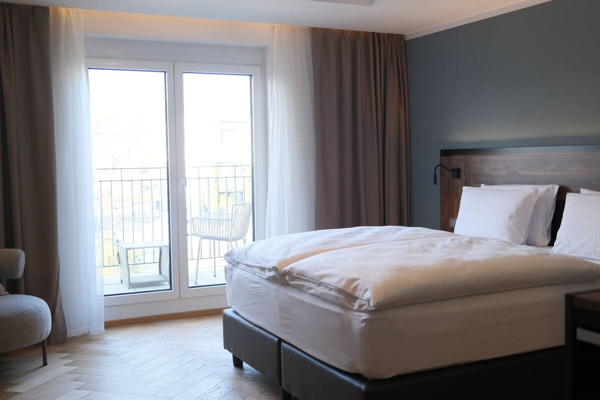 Hotellihuone, jossa on suuri sänky, puinen pääty, pieni yöpöytä ja näkymä parvekkeelle, jossa on ulkokalusteita.