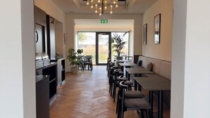 A modern interior with a herringbone wooden floor, a central seating area with tables and chairs, a kitchen area with a grill, and a ceiling with a geometric light fixture. - UNICO Boutique Hotel (Hochheim am Main)