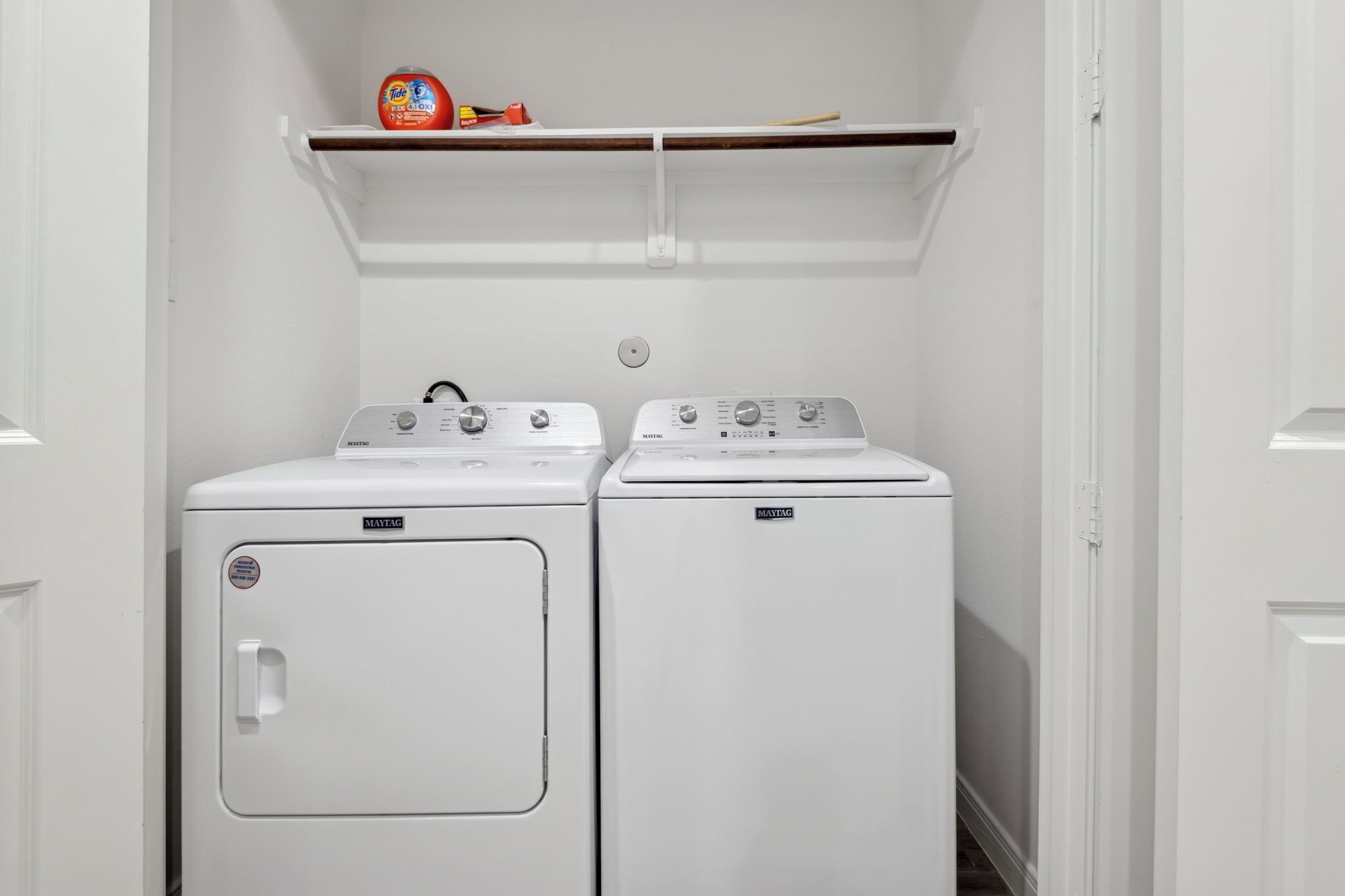 A laundry room with two Maytag front-loading washers and a shelf above holding laundry supplies.