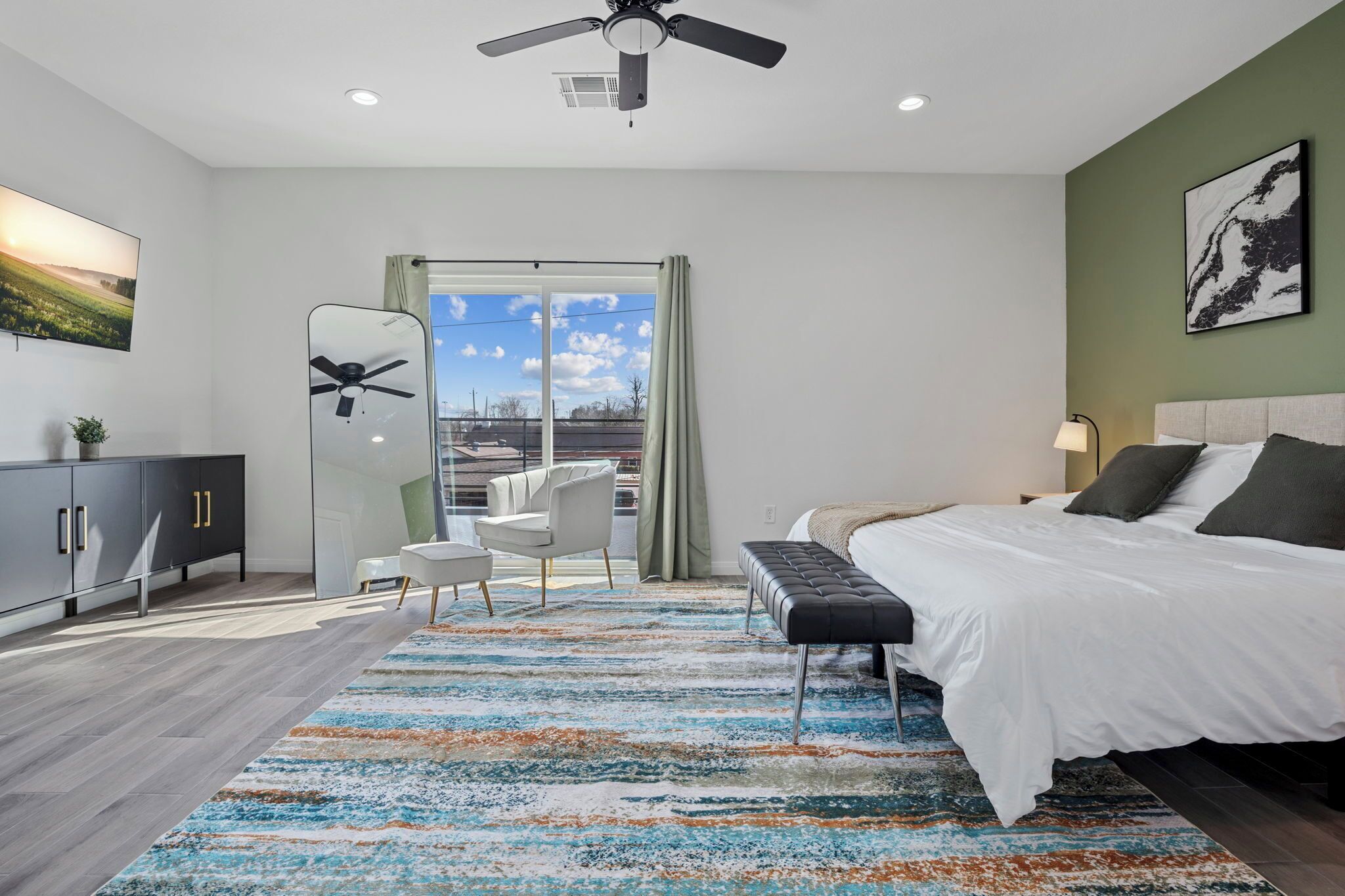 A modern bedroom with a large bed, a desk, a chair, and a view of the outdoors through a sliding door.