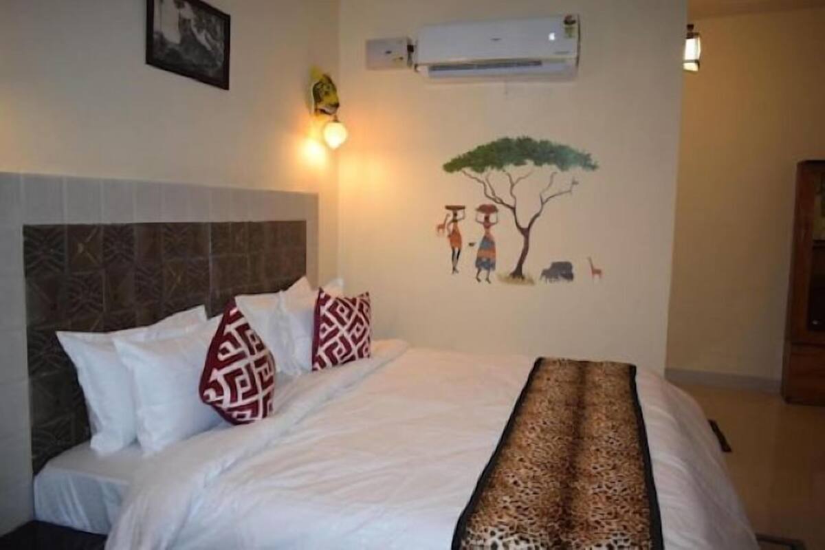 A neatly made bed with patterned pillows, a wall-mounted air conditioner, and a wall art depicting two figures under a tree.