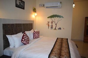 A neatly made bed with patterned pillows, a wall-mounted air conditioner, and a wall art depicting two figures under a tree. - Hotel Rann meadows (Sawai Madhopur)