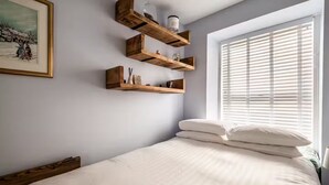Une chambre à coucher avec un lit, du linge de lit blanc, des étagères en bois ornées d’objets décoratifs, un tableau encadré au mur et une fenêtre avec des stores.