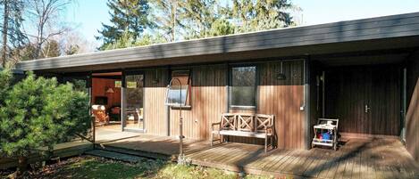 Una cabaña moderna con terraza de madera, un porche cubierto y un amplio ventanal con vistas a un bosque.