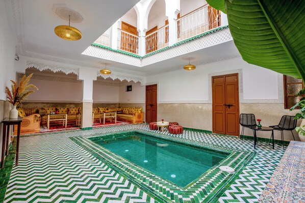 A spacious indoor swimming pool with a geometric tile floor, a seating area with patterned cushions, and a balcony with a railing. - Riad Ilyan & Spa  (Marrakech)