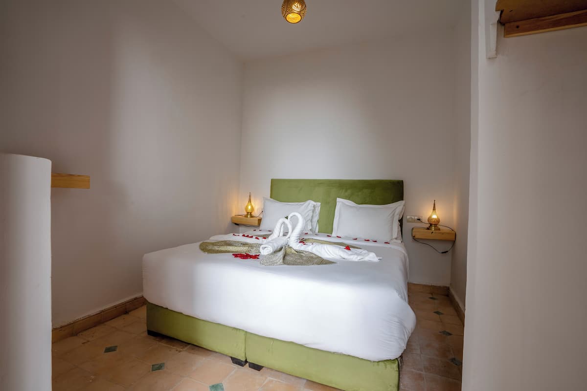 Una cama bien tendida con sábanas blancas y un cabecero verde, dos lámparas de noche y un arreglo decorativo con cisnes en la cama.