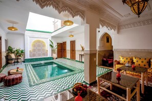 A spacious indoor area with a swimming pool, a seating area with a sofa and table, and a decorative ceiling with intricate patterns. - Riad Ilyan & Spa  (Marrakech)