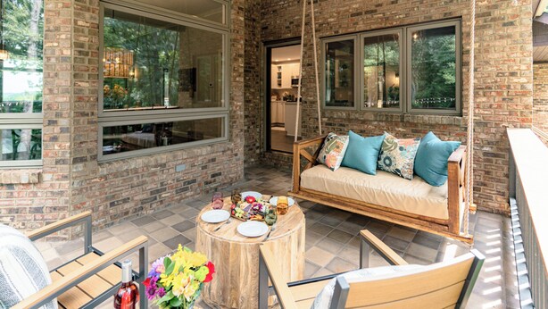 Un espace extérieur accueillant avec des sièges confortables, une balançoire, une table ronde en bois avec des assiettes et un vase rempli de fleurs, et un mur de briques agrémenté de grandes fenêtres.