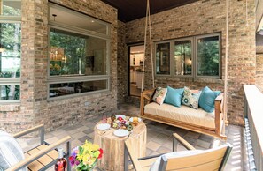 A cozy outdoor seating area with a swing, a round wooden table with plates and a vase of flowers, and a brick wall with large windows. - Rickenbacker Ridge sleeps 13 w/ heated pool+hot tub+firepit+game room ! (Sugar Grove)